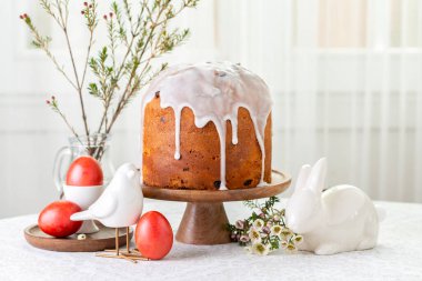 Easter food composition. Cake, sweet bread decorated with icing, red coloured eggs, spring flowers, table and window background. Easter holiday symbols, bird and bunny. White background.
