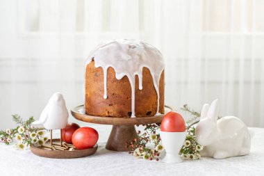Easter cake or sweet bread decorated with white icing, red coloured Paschal eggs dyed, spring flowers, table and window background. Easter holiday decorations and symbols: bird and bunny.