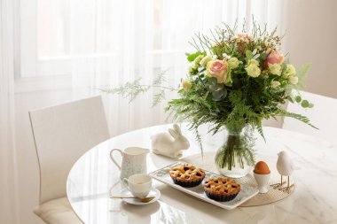 Beautiful morning breakfast table setting, chears. Flowers, espresso coffee, pastry, mini tarts or pies, hard boiled egg. White interior background with window. Spring or easter concept. 