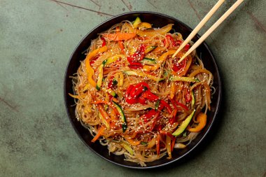 Top view of a plate with Japchae , Korean glass noodle made with sweet potato starch,  stir fried with vegetables.  Zucchini, bell pepper, carrot, sesame seed. Green table surface.