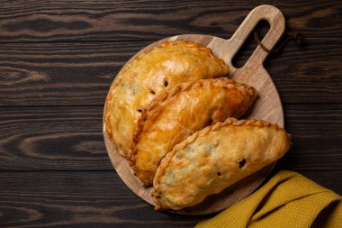 Flat lay of baked meat turnovers or pies, or empanadas, or cornish pasty with filling, beef, carrot, and potato on a dark wooden board.