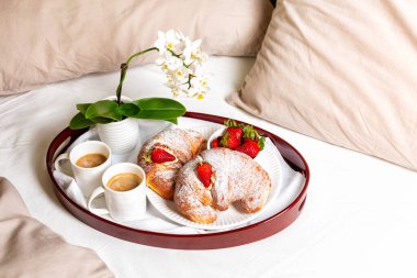 Yatak odasının yatağında kahvaltı. Bir çift için iki fincan kahve, kremalı ve çilekli kruvasan. Beyaz çiçekler. Romantik konsept.
