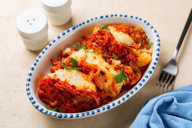 Served Greek and Polish style oven baked cod fish Psari Plaki with sauteed, shredded vegetables, carrot, onion, parsley and tomato sauce. White plate with blue decoration.	