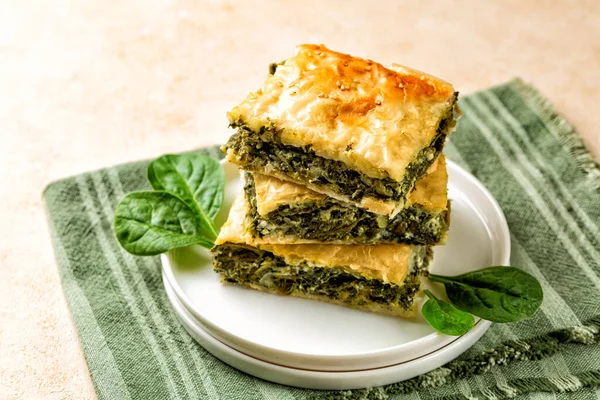 Stack of Spanakopita piece of pie, homemade Greek savory spinach pastry. It contains cheese feta, chopped spinach, green, egg, layered in phyllo or filo dought. Light beige background with green napkin.