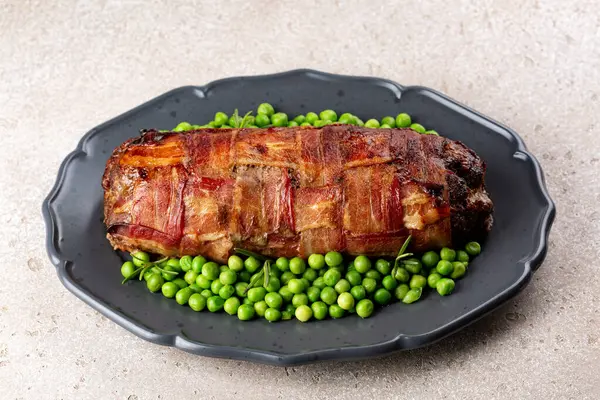 Baked meatloaf made of ground meat, onion, carrot and egg, wrapped with bacon net, served with green peas. Meat loaf is a traditional German, Scandinavian and Belgian dish.