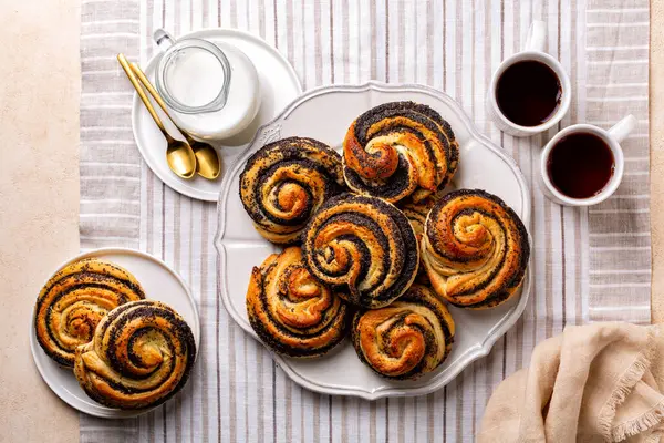 Top view of baked homemade poppy seeds buns, eastern European classic sweet yeast dough, swirl shape.  Milk and cup of tea. Breakfast on the table with linen white and beige runner.