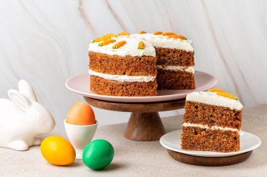 Paskalya kompozisyonu. Bir dilim havuçlu kek, renkli yumurtalar ve beyaz tavşan. Tatil yemeği, hafif arka plan..