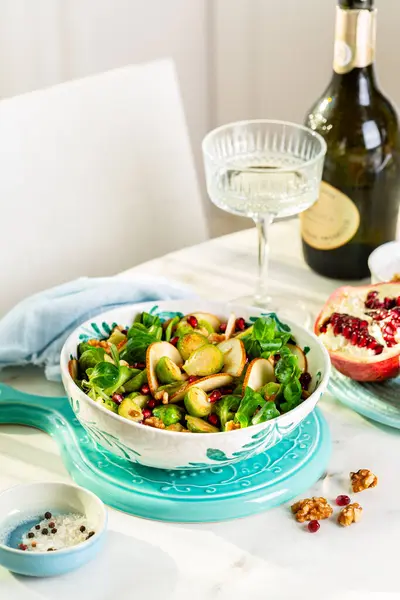 White Table with winter salad with roasted brussel sprouts,  pear, walnut, pomegranate, valerian salad. Ingredients. Wine glass and bottle. Vertical image.