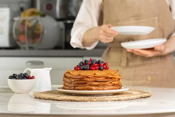 stock image Stack of sweet homemade crepes, type of thin pancake, with blueberry and red currant in the center of table. Woman in apron on background preparing kitchen table for breakfast, puttting plates. 