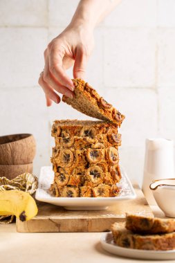 Bir dilim hindistan cevizi unlu muz ekmeği alıyorum. Kadın eli. Bej masa yüzeyindeki malzemeler. Dikey resim.