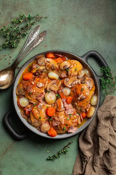 Coq au vin veya horoz, şaraplı horoz. Kırmızı şarap, lardon, mantar, havuç ve soğanla pişirilmiş Fransız tavuğu. Tuz, biber ve kekik ile tatlandırılmış. Yeşil masa yüzeyi. Dikey resim.