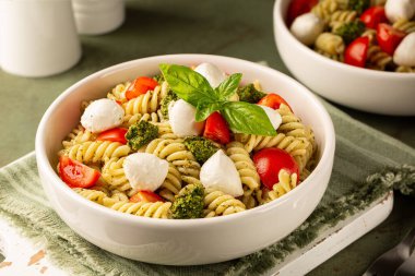 Plate with cold pasta salad with homemade pesto sauce, tomato and mozzarella cheese, decorated with basil leaves on a green table surface. Italian lunch. clipart