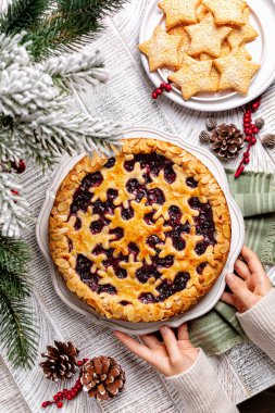 Winter Christmas or New Year festive bakery. Hands holding baked mixed berry almond crust pie or tart with snawflakes decoration. Homemade star shaped cookies on a white table, vertical image. clipart