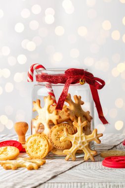 Ev yapımı kar tanesi şekilli ve yuvarlak şekilli kurabiyeler Mutlu Yıllar ve Mutlu Noeller, kırmızı kurdeleli cam kavanoz, ışıklarla beyaz zemin, dikey görüntü.