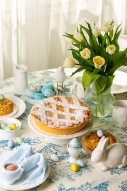 İçten servis edilen paskalya yemeği masası, İtalyan paskalya pastası, ricotta peynirli kısa kabuklu turta, Pastiera Napoletana, renkli yumurtalar, çörekler, paskalya tavşanı ve kuş heykelleri, lale çiçekli vazo. Dikey resim.