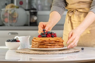 Önlüklü kadın ev yapımı krep yığınına böğürtlen koyuyor. Masanın ortasında yaban mersini ve kırmızı üzümlü krep var.. 