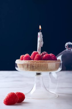 Kremalı tart ve taze olgun ahududu ile siyah arka planda ışık mumu, doğum günü ve kutlama konsepti.