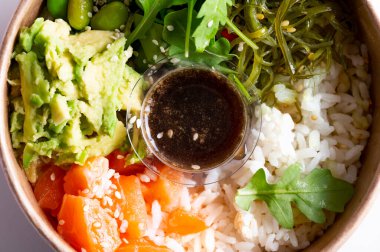 Suşi pilavı, çiğ somon, avokado, yosun salatası, roket, edamame fasulyesi ve taze soğanlı soya susamlı soslu paket kâğıt kâsesi.