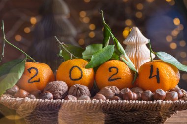 Yeni Yıl 2024 konsepti geliyor. Siyah mürekkeple yazılmış rakamlar mandalinalar, sepetin içinde fındıklı portakallar ve arka planda Xmas ışıkları.