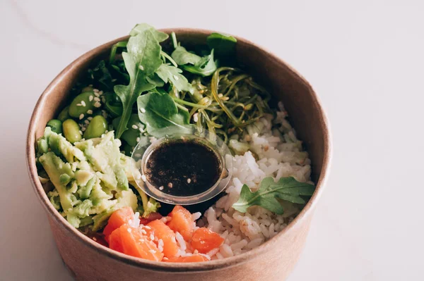 stock image Poke bowl with sushi rice, raw salmon, avocado, seaweed salad, rocket, edamame beans and spring onion with a soy sesame dressing in take away paper bowl