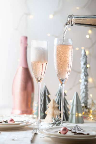 Xmas table settings in white and pink colors with trendy paper christmas trees on background. Table ready for festive dinner and party with pink prosecco pouring to the glass