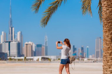 Dubai arka planında çekimler yapan turist bir kadın. Modern şehir merkezi, BAE 'de gökdelenleri var. Ünlü seyahat noktasında güzel bir model gezisi. Palmiye ağaçları ve güneşli bir günü olan güzel kumsal.