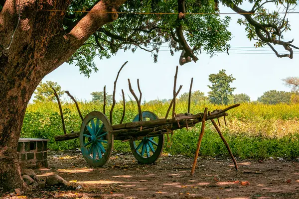 stock image A vintage wooden cart rests idyllically under the shade of a lush tree in a tranquil rural setting
