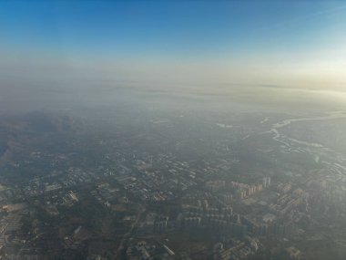 Yukarıdan puslu bir atmosferi olan bir şehir görülüyor. Gökyüzü mavi ve şehir binalarla dolu.