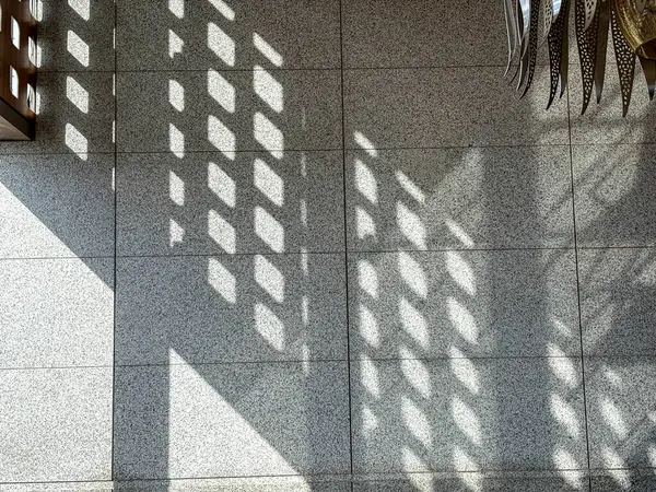 A shadow of a tiled window is cast on a tiled floor. The shadow is large and dark, and it covers a significant portion of the floor