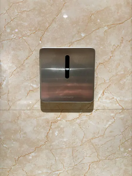 A silver coin slot urinal human detector on the wall is tiled and the tile is beige