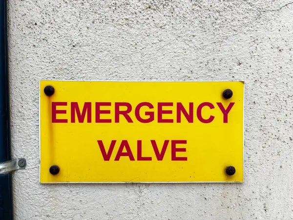 Front view of a yellow sign board displayed on a cream color wall saying \'Emergency valve\'. The text written in red colour. A black pole is running vertically the on the side of the board.