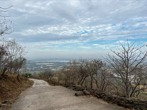 A path winds through a forest with a view of a city in the distance at Ralamandal, Indore, India.. The sky is cloudy, but the sun is still visible.