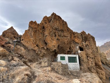 Küçük bir ev büyük bir kayanın içine inşa edilir. Ev yeşil ve penceresi var. Kaya kahverengi ve gökyüzü bulutlu.