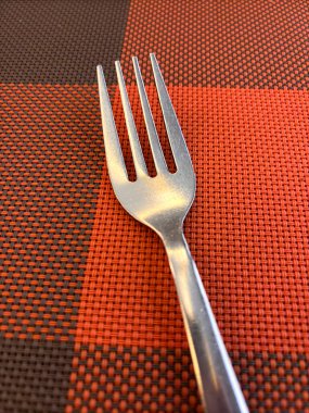 Bir çatal kareli bir masa örtüsünün üzerinde oturuyor. Çatal gümüşten ve üç çatalı var. Masa örtüsü turuncu ve kırmızıdır. Sıcak ve davetkar bir atmosfer yaratır.