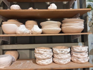 Beyaz seramik kaseler ve vazolarla dolu bir raf. Kaseler çeşitli boyutlarda ve şekillerde, vazolar uzun ve incedir. Sahne, yaratıcılık ve işçiliğin bir parçasıdır.