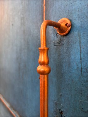 Altın saplı metal bir boru bir duvara bağlıdır. Boru eğilmiş ve dekoratif bir tasarımı var. Duvar mavi ve boru turuncu.