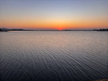 Güneş sakin bir su kütlesinin üzerinde batıyor. Gökyüzü turuncu ve mor renklerin bir karışımıdır. Huzurlu ve huzurlu bir atmosfer yaratır. Su durgun, dalgalanma veya dalgalanma yok.