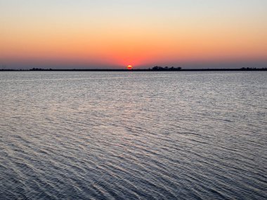Güneş sakin bir su kütlesinin üzerinde batıyor. Gökyüzü turuncu ve mor renklerin bir karışımıdır. Huzurlu ve huzurlu bir atmosfer yaratır. Su durgun, dalgalanma veya dalgalanma yok.