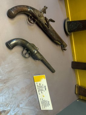 Sarı arka planı olan bir masada iki eski silah sergileniyor. Silahlar eski ve paslı ve masa sarı bir bezle kaplı. Manzara nostaljik ve ürkütücü.