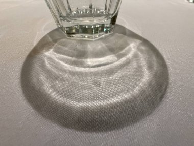 Bir masanın üzerinde bir bardak oturuyor ve onun gölgesi masanın üzerine düşüyor. Gölge büyük ve karanlık, derinlik ve boyut hissi yaratıyor. Bardağın kendisi berrak ve dikdörtgen.