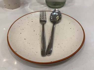 Gümüş bir çatal ve kaşık, kahverengi kenarlı beyaz bir tabakta. Tabak bir masaya konur.