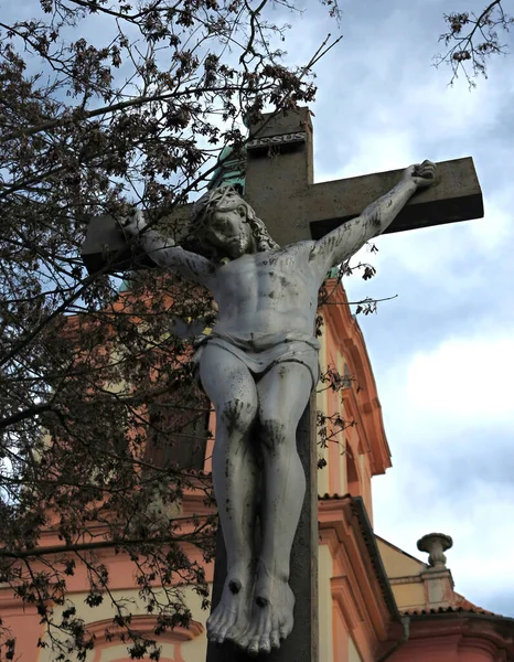 Statue of martyred Jesus on the cross by the church. Silver retro statue of Jesus under the shadows of a tree on a wooden cross. City capital city of Prague.