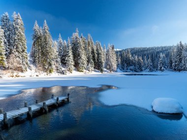 Dağlarda karlı bir arazide eski bir ahşap iskelesi olan güzel bir göl manzarası. Eriyen göl, gökyüzünde bulutlu güneşli hava. Duvar kağıdı, arkaplan.