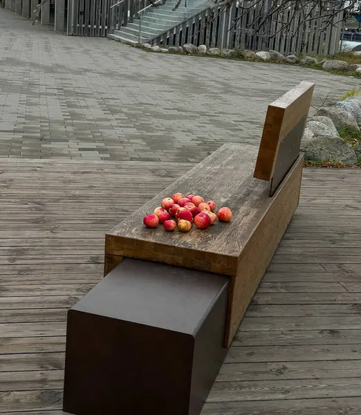Bir grup kırmızı elma, ahşap bir bankta modern metal detaylarla yerleştirilmiş, ahşap bir döşeme ve taş bir patika ile kentsel bir ortamda fotoğraflanmış.. 