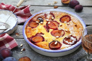 Damson blue plum clafoutis (flan) and glass of rose wine, icing sugar and almonds dressing, french cuisine on wooden background. Rustic style