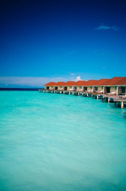 Güneşli bir günde turkuaz okyanusun üzerinde kırmızı çatıları olan Overwater Resort bungalovları. Maldivler 'de berrak gökyüzü ve sakin denizle lüks tropikal tatil sahnesi