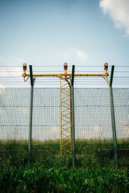 Havaalanı güvenlik çiti yeşil tel örgü ve sarı metal kule yapısının arkasında pist ışıkları var..