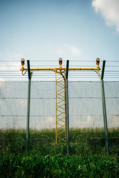 Havaalanı güvenlik çiti yeşil tel örgü ve sarı metal kule yapısının arkasında pist ışıkları var..
