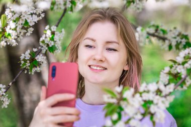 Mor tişörtlü bir kadın arkadaşlarıyla görüntülü konuşma yapıyor. Elma bahçesinde beyaz çiçekli yumuşak bir fotoğraf. Akıllı telefon ve teknoloji konsepti.