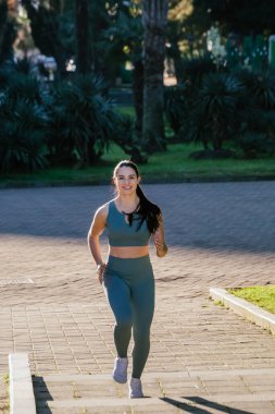 Sportif ve güler yüzlü genç bayan spor giyim koçu parkta koşuyor. Spor açık hava konsepti.